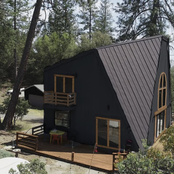 A-frame cabin with dark exterior, wooden deck, and large arched window, surrounded by trees and greenery.