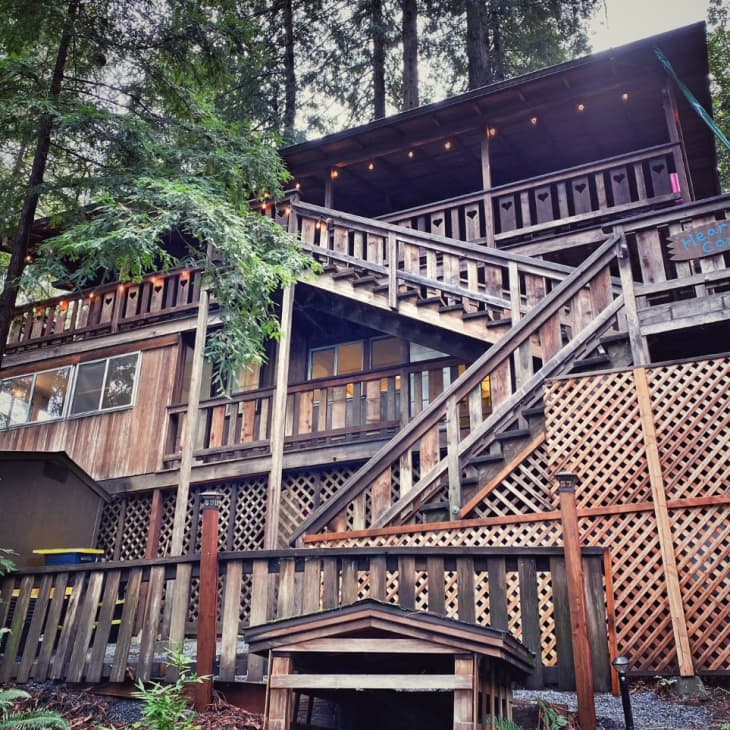 Rustic multi-level wooden cabin with latticework and surrounded by tall trees.