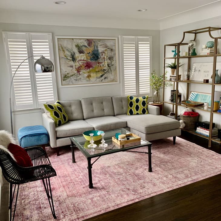 Modern living room with gray sectional sofa, colorful pillows, abstract art, and a gold bookshelf on a pink rug.