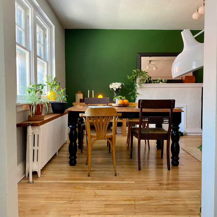 Dining room with wooden table, mixed chairs, green accent wall, plants, and a large white pendant light.