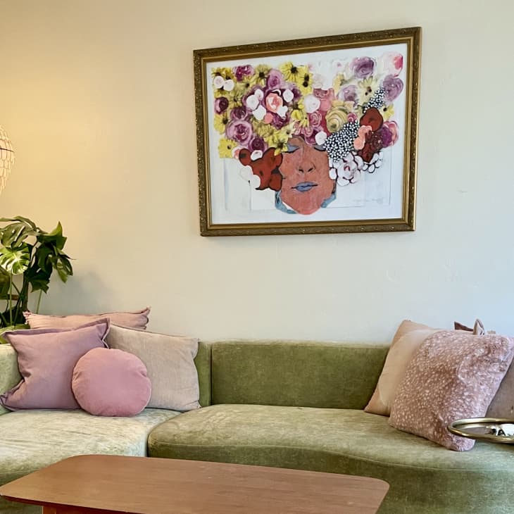 Green velvet sofa with pink cushions, abstract floral artwork, pendant light, and potted plant in a cozy living room.