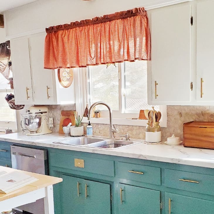 Kitchen with teal lower cabinets, white upper cabinets, marble countertops, red curtains, and a mixer on the counter.
