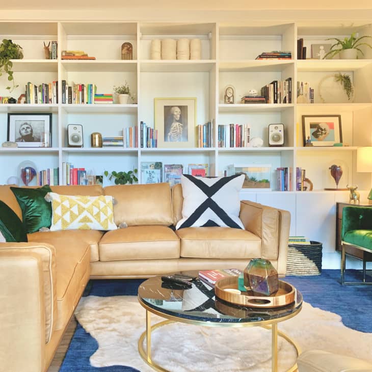 Beige colored living room with bookshelves along one wall, a camel-colored leather sectional sofa, and velvet green side lounge chairs.
