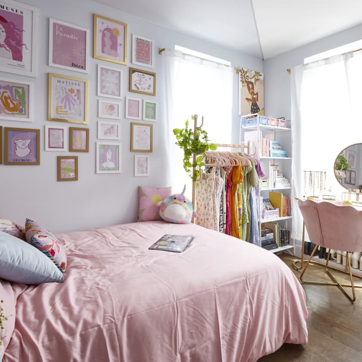 Bedroom with pink bedding, gallery wall, vanity with round mirror, and clothing rack by a window with sheer curtains.