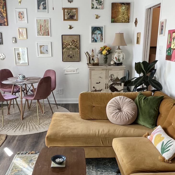 Living room with mustard velvet sofa, pink and green pillows, round dining table with pink chairs, and eclectic wall art.