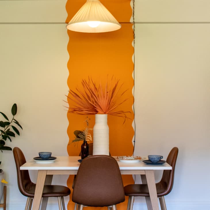 Dining area with white table, brown chairs, orange accent wall, pendant light, and potted plants.