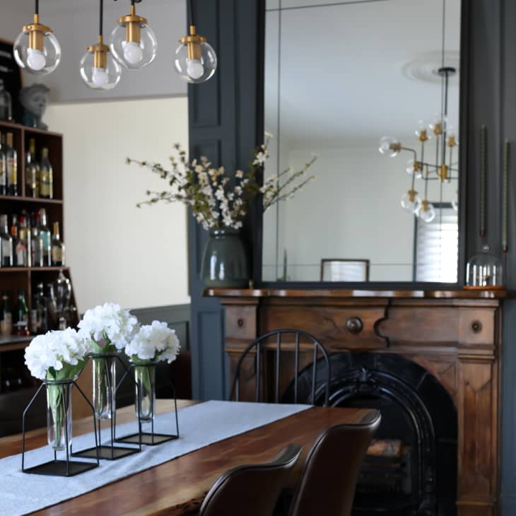 Dining room with wooden table, white flowers in vases, large mirror, and a bar shelf with bottles.