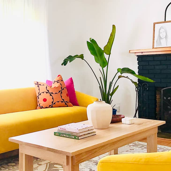 Bright living room with yellow sofas, colorful pillows, wooden coffee table, potted plant, and black fireplace.