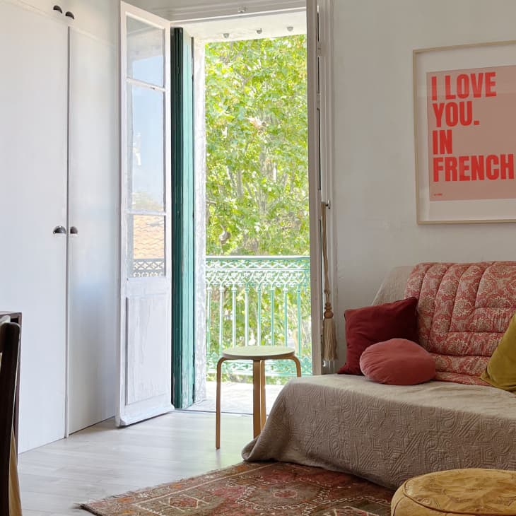 Cozy living room with a patterned sofa, colorful cushions, wall art, and open doors leading to a balcony with greenery.