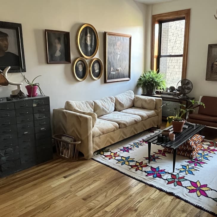 Living room with tan vintage patterned sofa, star pattern flatweave rug, slatted black coffee table, apothecary style drawer black dresser, framed portraits on sofa wall