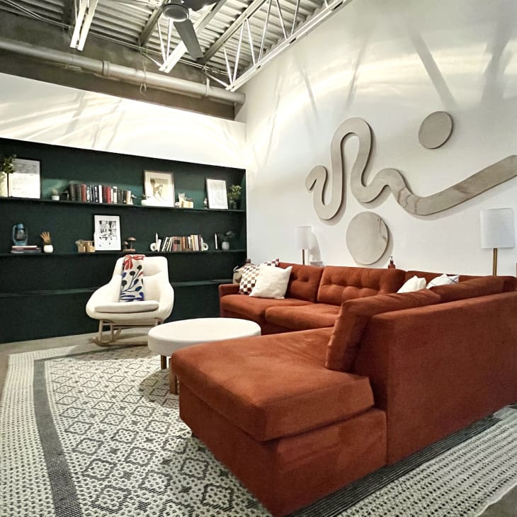 Living room with white walls, burnt orange/rust colored velvet sectional sofa, white and gray patterned rug, white round coffee table and white accent chair, large black bookshelves on back wall, geometric type art above sofa