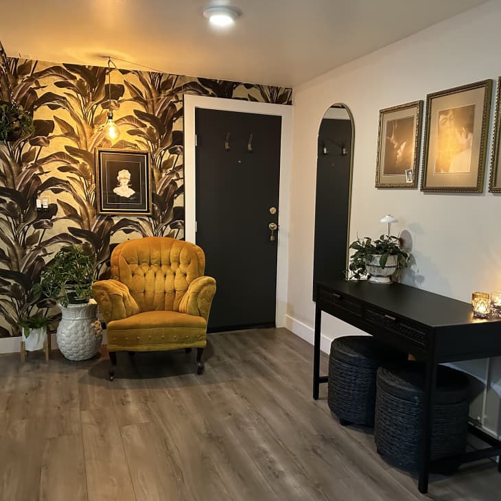 Apartment with lots of neutral, natural hues, Italian art, natural and black accents, plant=patterned wallpaper. This is a seating  area with a gold plush chair, black accent table with 2 black ottomans