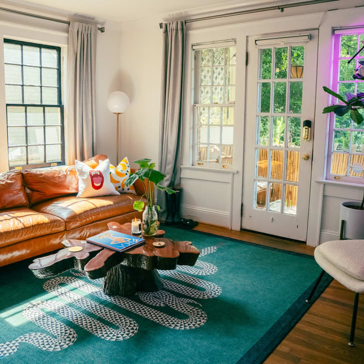 living room with camel leather sofa, teal rug with snake pattern, tree-trunk shape coffee table, plants, lots of paned windows and door, white walls, white leather accent chair with ottoman, wood floor