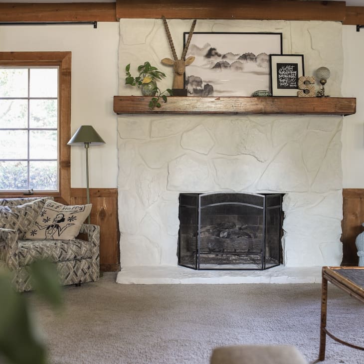 white stone fireplace, natural wood mantle, wood paneling half wall, beige carpet, green curtains, wood window trim and crown molding, green floor lamp, art, deer head sculpture, bamboo coffee table, vintage patterned arm chairs, white and brown arm chairs, tri leg lamp, plants