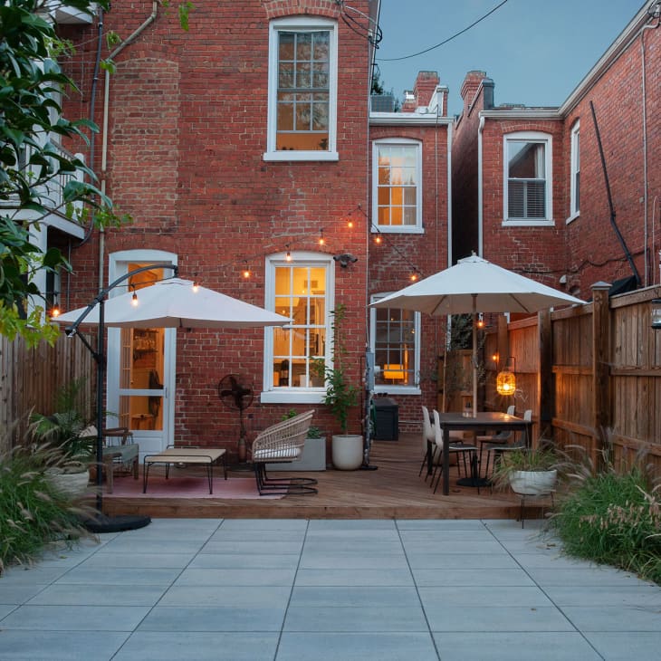 Back of brick building with patio dining area with white umbrellas, cement driveway, wood fences, string lights