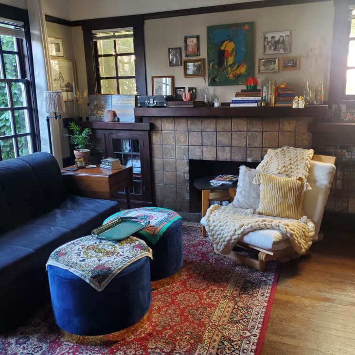 Cozy living room with dark sofa, blue ottomans, patterned rug, and a fireplace adorned with books and framed art.