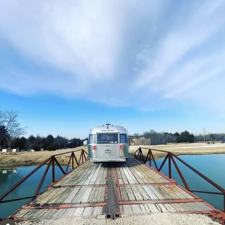 Airstream trailer on a wooden bridge over a pond, surrounded by grassy fields and trees under a blue sky.