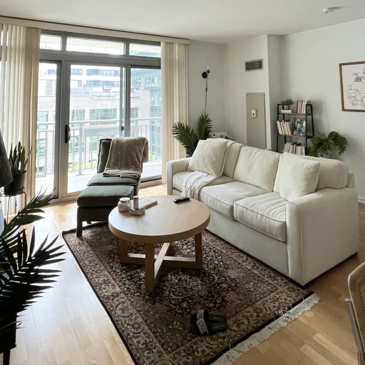 Living room with white walls, white sofa, wood floor, large sliding glass doors with cream vertical blinds, brown Persian area rug with cream fringe, round wood coffee table, part of dining area can be seen in foreground