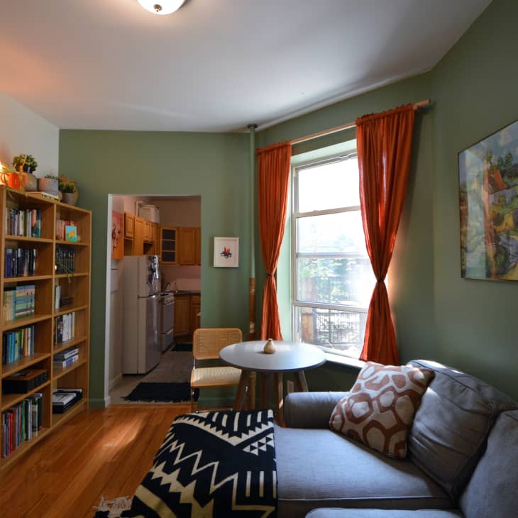 natural wood book shelf, orange curtains, grey couch, black and white geometric pattern rug, green walls, landscape art, throw pillow, door way to kitchen, wood floors