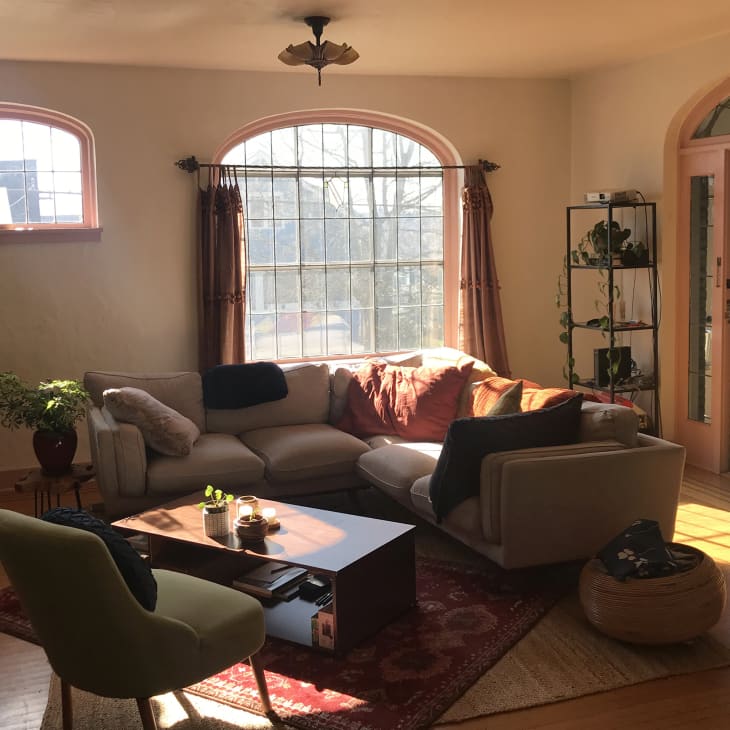Living room with warn sunlight coming in, pale gray sofa with orange, green, blue throw/pillows, arched window with blush trim, curtains. Blush trim on arched door, shelves with plants, wood floor, wood coffee table with accent chair, tall plants to the left
