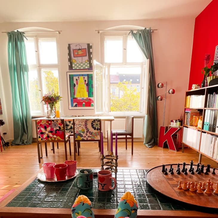 Colorful living room with red accents, floral chairs, bookshelves, chess set, and vibrant artwork on walls.