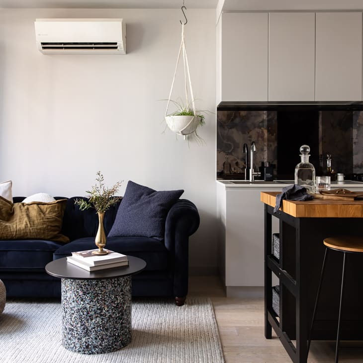 Living room and kitchen space in apartment with velvet blue sofa, small multi colored speckled coffee table and dark colored kitchen with wooden countertops.