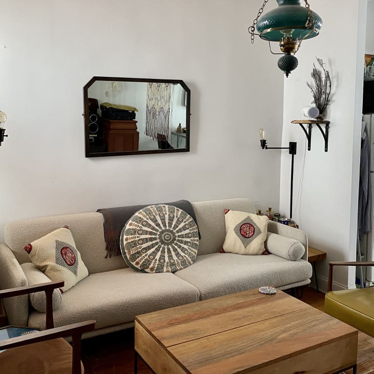 Living room with pale neutral sofa with patterned cream, black throw pillows, black framed mirror on wall behind, teal hanging lamp/lantern, greenish yellow leather chair, wood coffee table, wood armchair, wood floors, plants, white walls