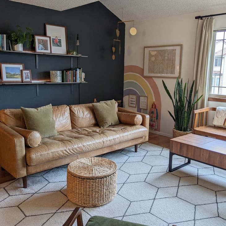 Living room with lots of neutral and natural tones. Brown leather sofa, pale neutral geometric pattern rug, wood bench with wood coffee table, straw ottoman basket. Navy blue wall behind sofa with shelves with books, art. Snake plant on floor next to wall rainbow mural