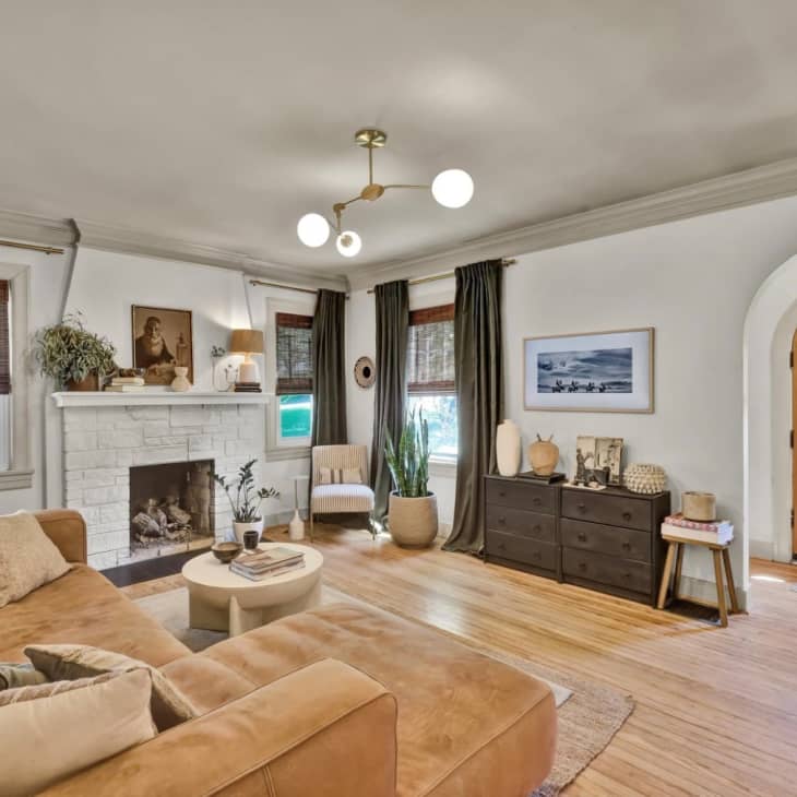 Living room with wood floors, arched front door, brown leather sofa, painted white brick fireplace, white walls
