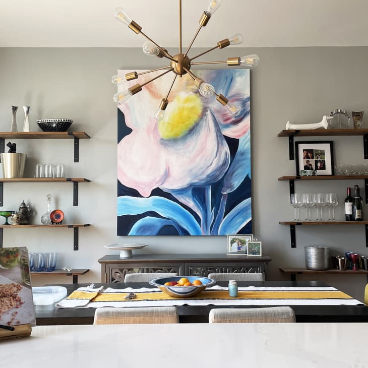 Dining room with long table with stripe runner, large flower painting, wood wall shelves with glasses, barware, art objects, sputnik style pendant light