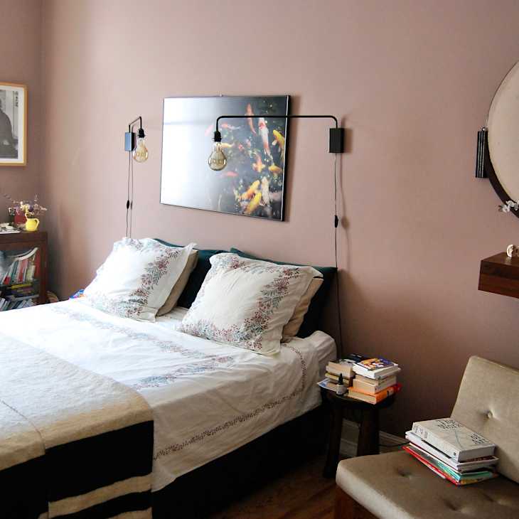 Bed with floral print bedding with hanging bulb lamps over it, stacks of books on nightstand and chair, and dusty pink wall