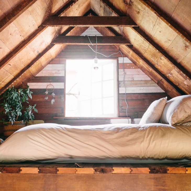 Cozy attic bedroom with wooden beams, a bed with beige linens, a potted plant, and a window with natural light.