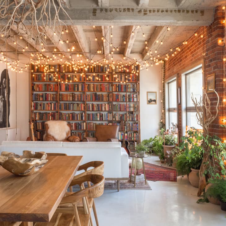 Loft living room with a wooden dining table, bookshelves, string lights, plants, and large windows.