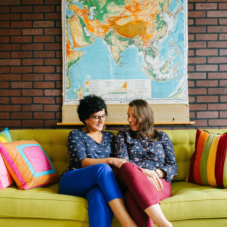 Two people sitting on a green sofa with colorful pillows, brick wall, and world map in the background.