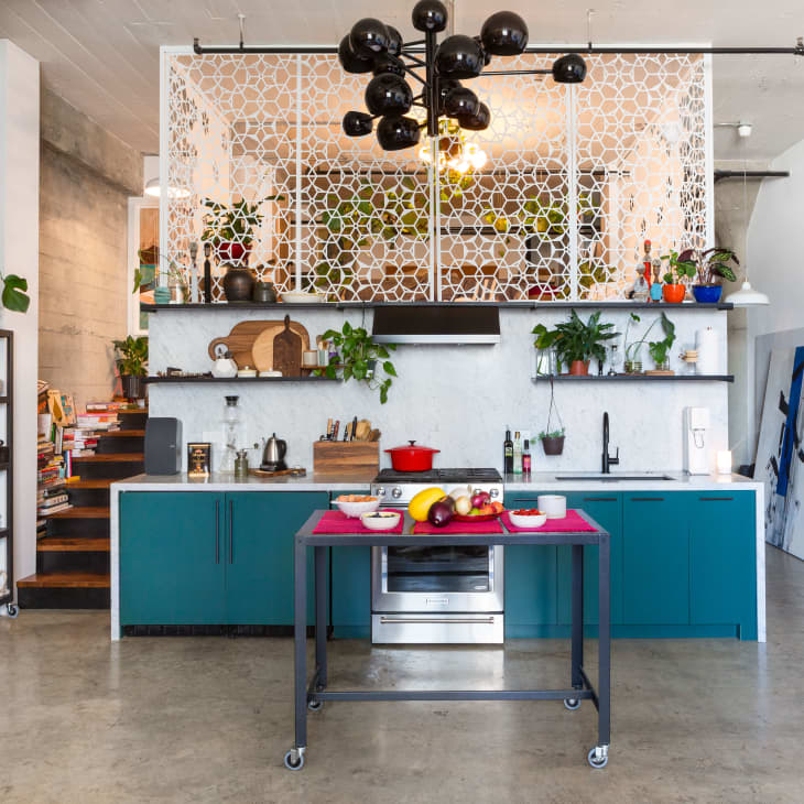 Eclectic kitchen with teal cabinets, open shelving, plants, and a decorative screen. Island with fruit and a red pot.