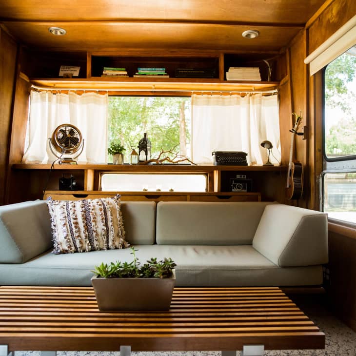 Mid-century modern trailer interior with a gray sofa, wooden coffee table, vintage fan, typewriter, and books on shelves.