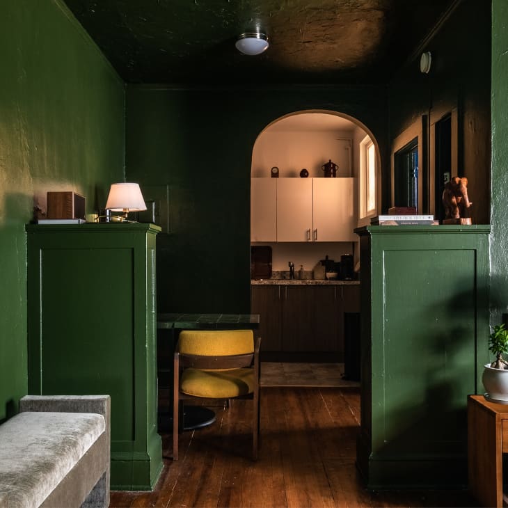 Green walls and ceiling, small half pillar room dividers, gray velvet bench, yellow mid century modern chair, small wood night stand, arched entrance to kitchen