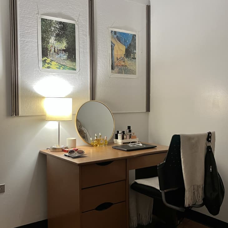 desk with small round vanity mirror and desk lamp, two impressionist paintings above desk and perfume on desk, white rolling chair