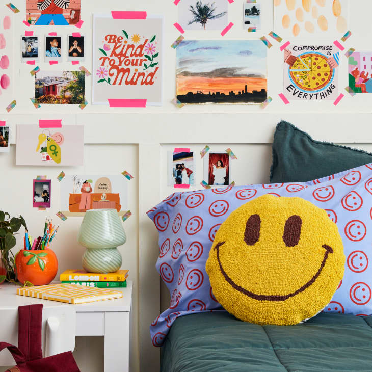 semi wide shot of a bed with teal sheets, blue pillowcase with red smiley faces on it, a smiley face pillow and a white desk with a red backpack hanging off white chair, a green lamp and supplies on top of desk, and a wall with different photos and art