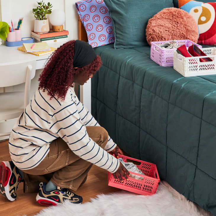 girl in dorm room crouching down placing a pink basket with shoes underneath bed