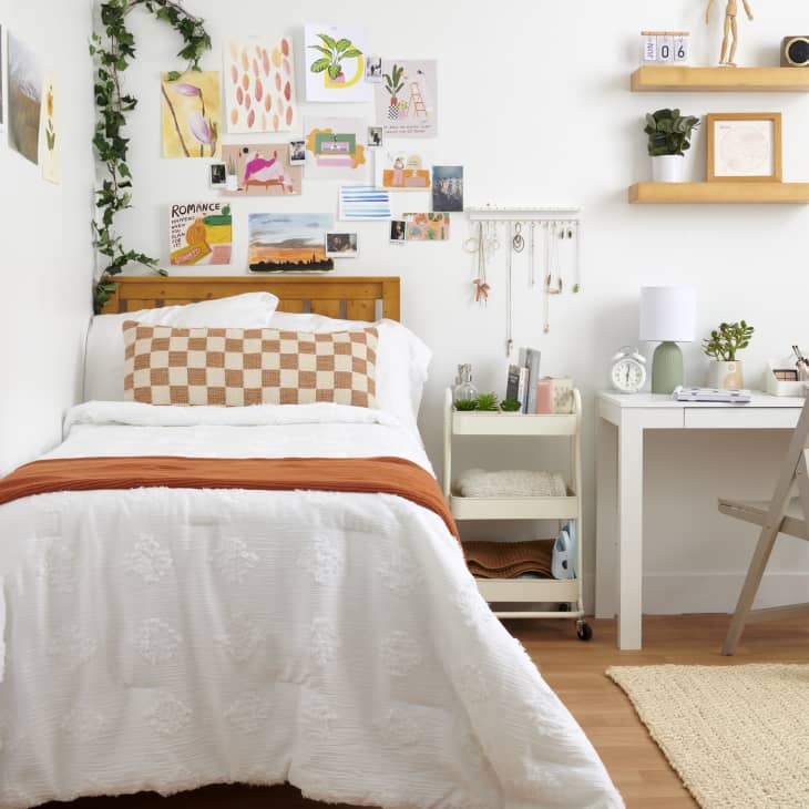 forward shot of bedroom where the bed has orange and cream checkered pillow and orange blanket, a gallery wall above the headboard - a rolling cart next to bed and a desk