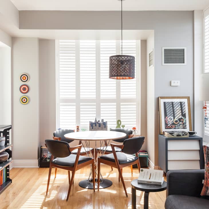 Modern dining area with round table, four chairs, pendant light, and wall art, next to a cozy living space with a colorful pillow.