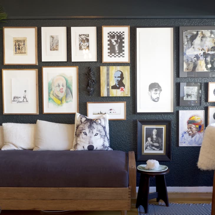 Gallery wall with eclectic art above a daybed, wolf pillow, and plants in a cozy living room setting.