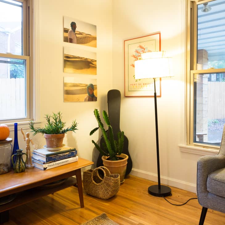 Cozy living room with a gray armchair, floor lamp, cactus, and wall art. Coffee table with books, plants, and a pumpkin.