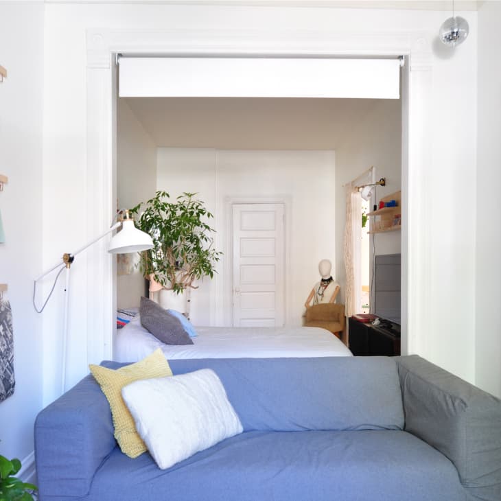Gray sofa with yellow and white pillows in a living room, leading to a bedroom with a bed, plants, and wall art.