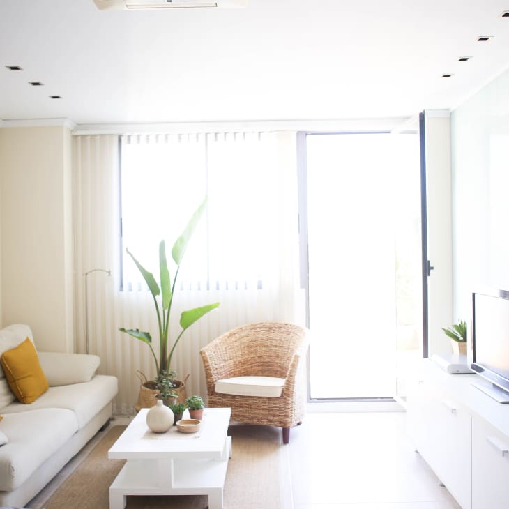 Bright living room with a beige sofa, yellow cushions, wicker chair, white coffee table, and TV on a white console.