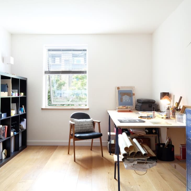 Home office with a black shelving unit, wooden chair, desk with art supplies, and a tall potted plant by the window.