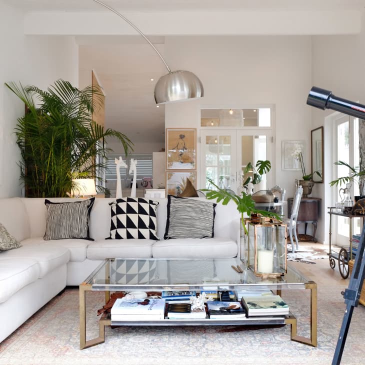 Modern living room with white sectional sofa, glass coffee table, large potted plants, and a telescope by the window.