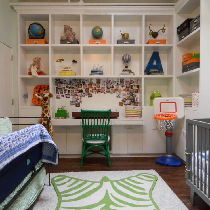 Child's room with a crib, green chair at a desk, wall shelves with toys, and a small basketball hoop.