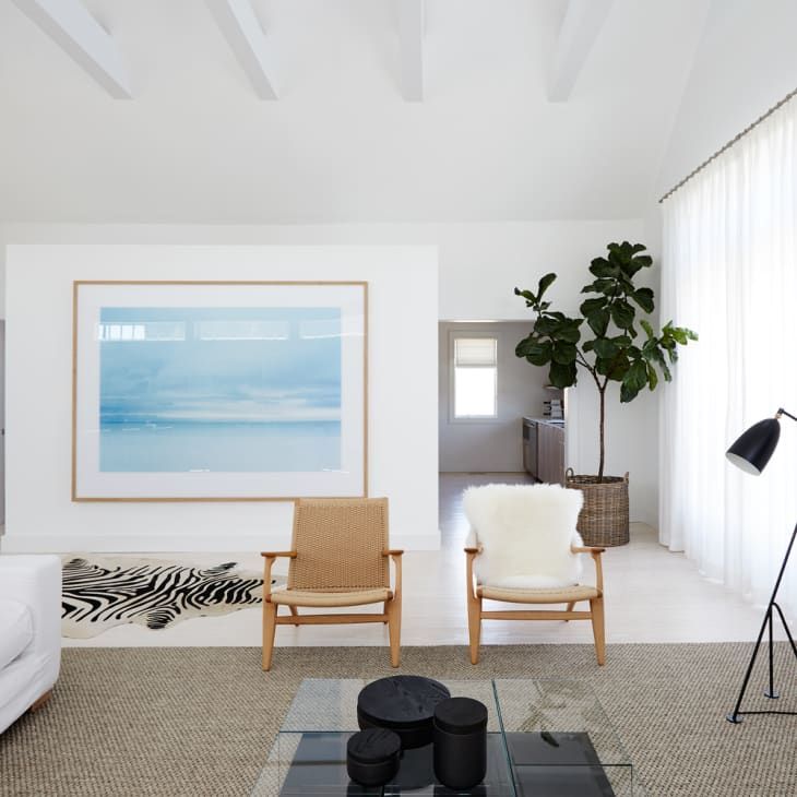 Modern living room with a white sofa, two wooden chairs, large framed ocean artwork, floor lamp, and a potted plant.